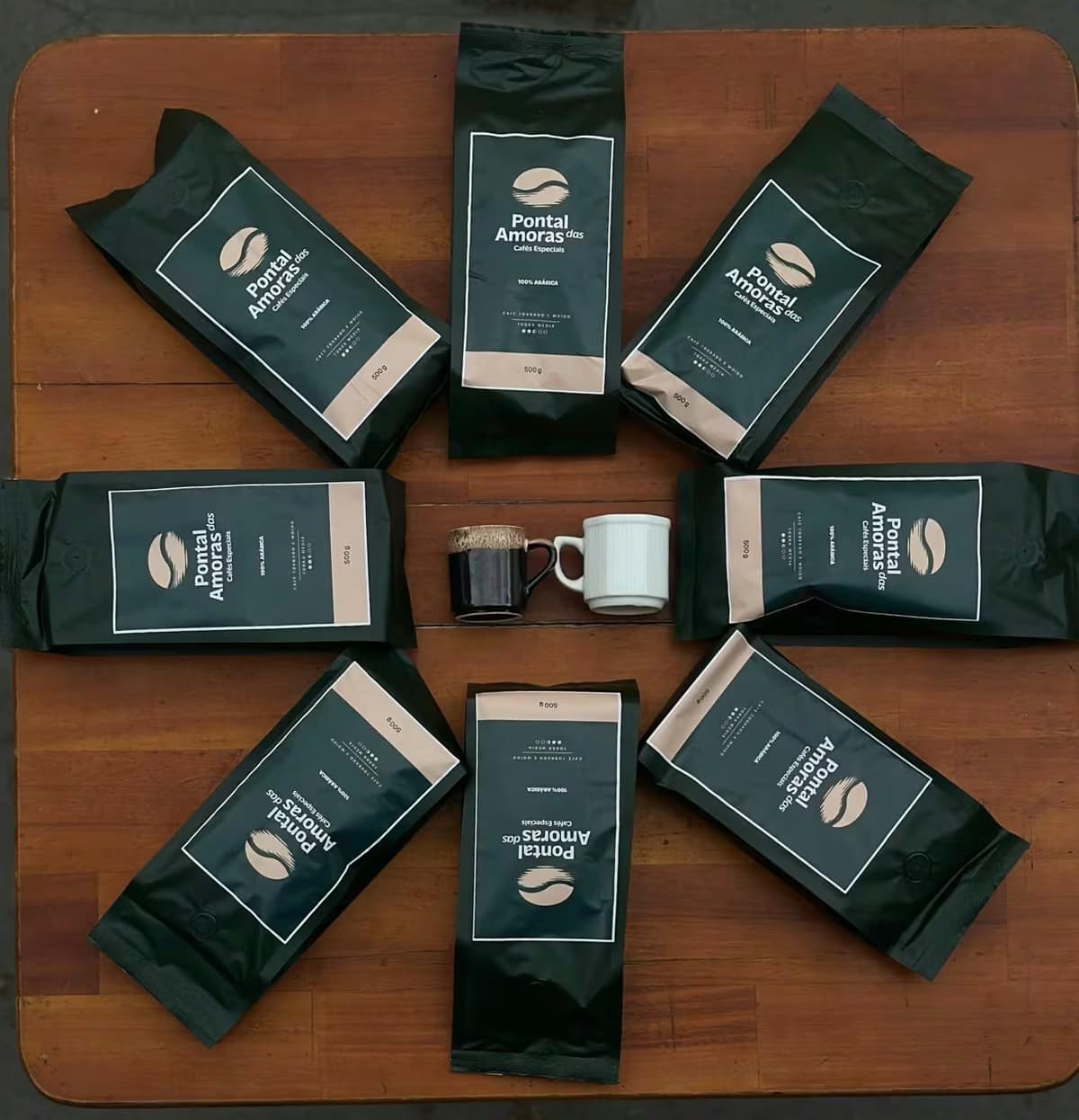 Oito pacotes de café Pontal das Amoras dispostos em círculo sobre mesa de madeira, com duas xícaras ao centro.
